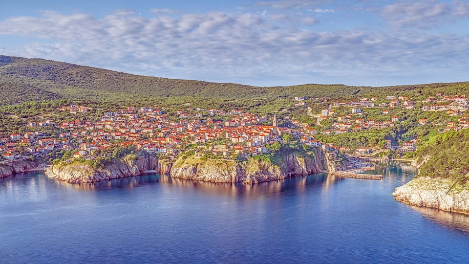 A coastal town nestled between hills, with a calm bay and dense greenery.