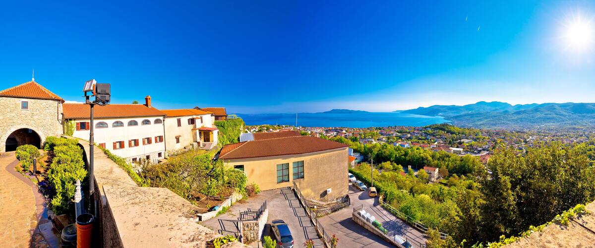 A coastal town with stone buildings, a paved road, and a view of the sea and mountains.