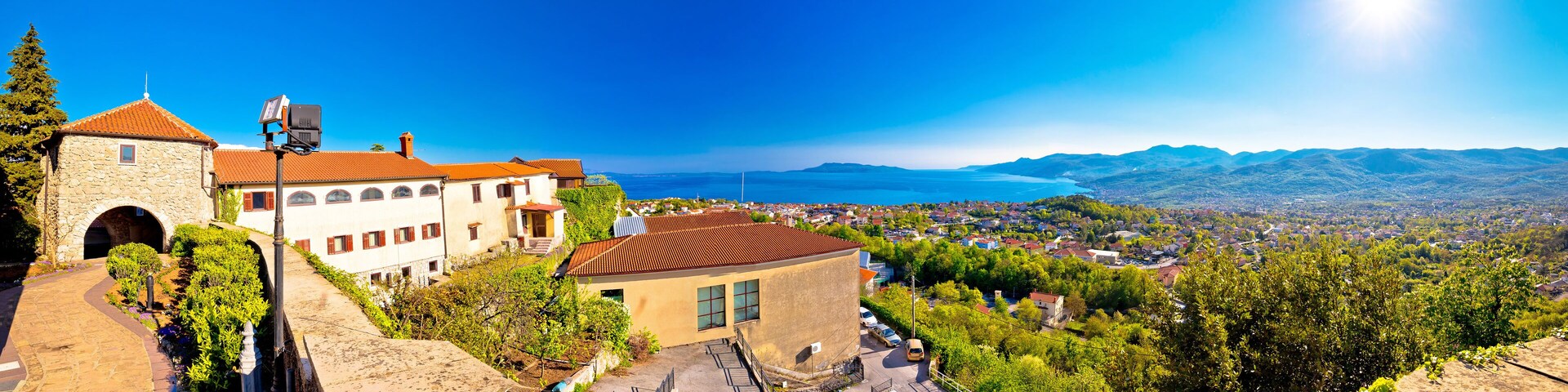 A coastal town with stone buildings, a paved road, and a view of the sea and mountains.