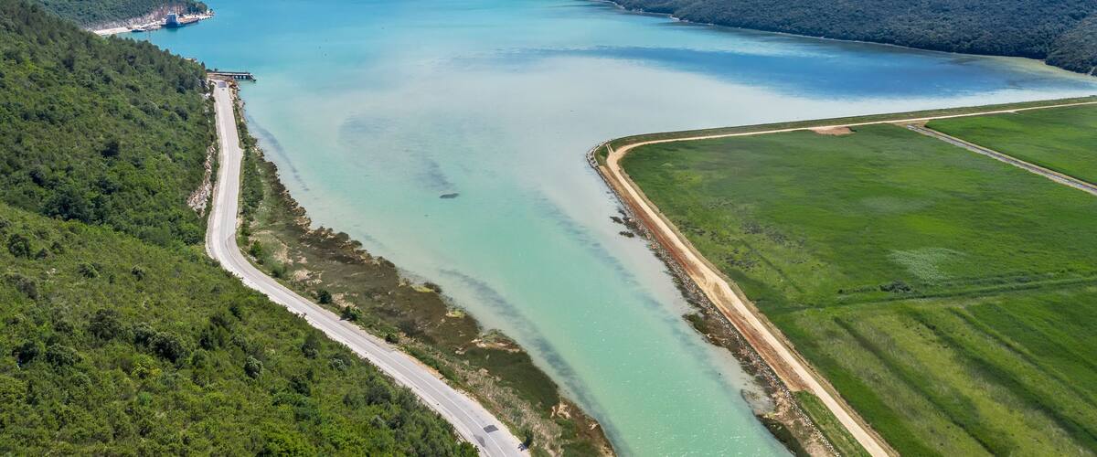 A coastal landscape with a clear blue lake, surrounded by green hills and a road leading to a small settlement.