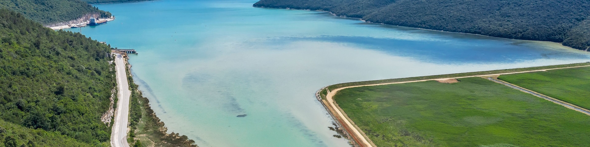 A coastal landscape with a clear blue lake, surrounded by green hills and a road leading to a small settlement.