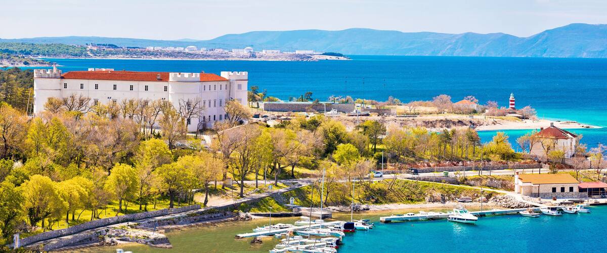 A coastal town with a castle, a lighthouse, and boats docked at the harbor.