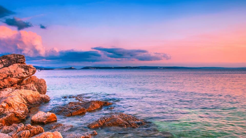 A rocky coastline at sunset with calm waters and distant landforms.