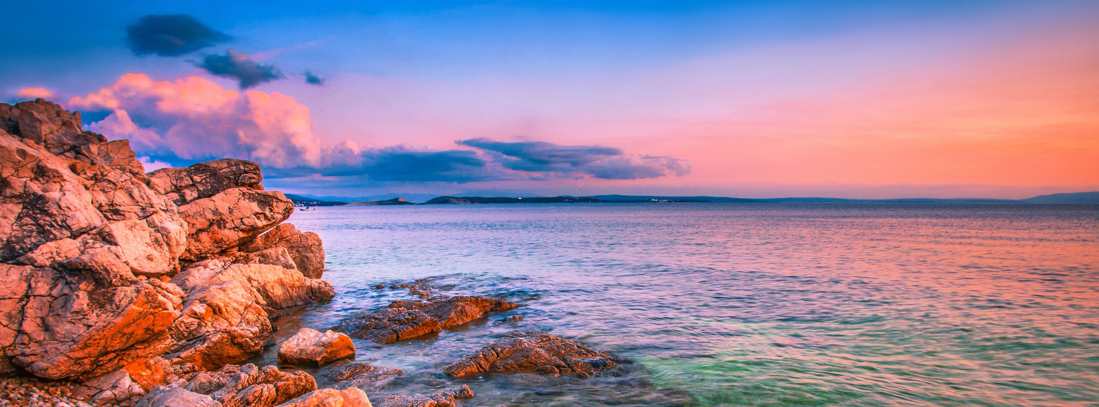 A rocky coastline at sunset with calm waters and distant landforms.