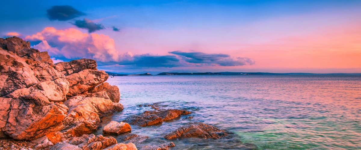 A rocky coastline at sunset with calm waters and distant landforms.