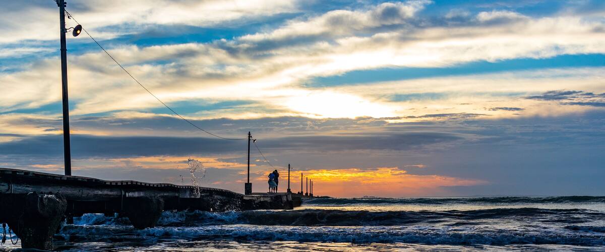 A sunset over a beach with waves, a pier, and a person standing on the pier.