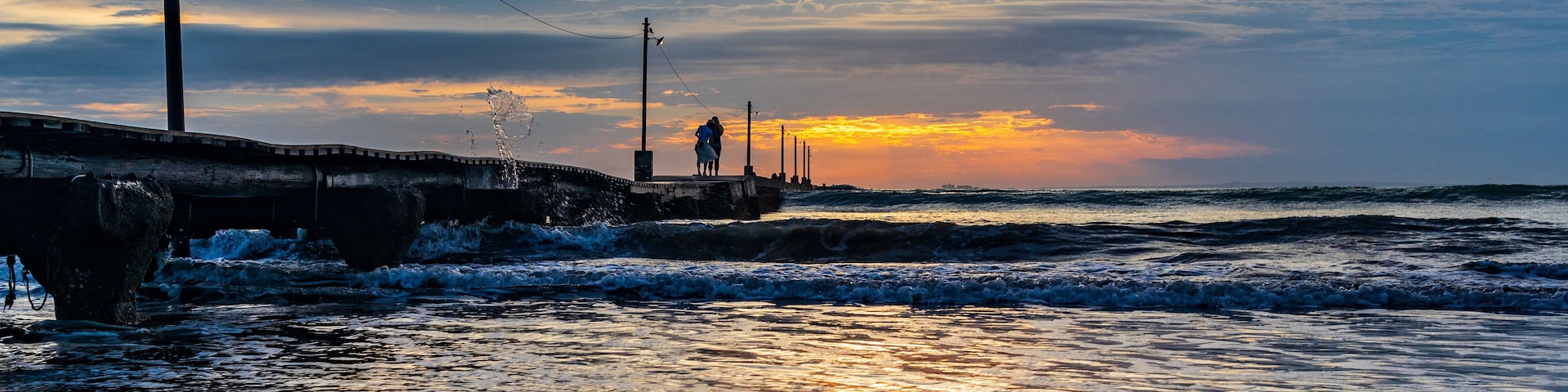 A sunset over a beach with waves, a pier, and a person standing on the pier.