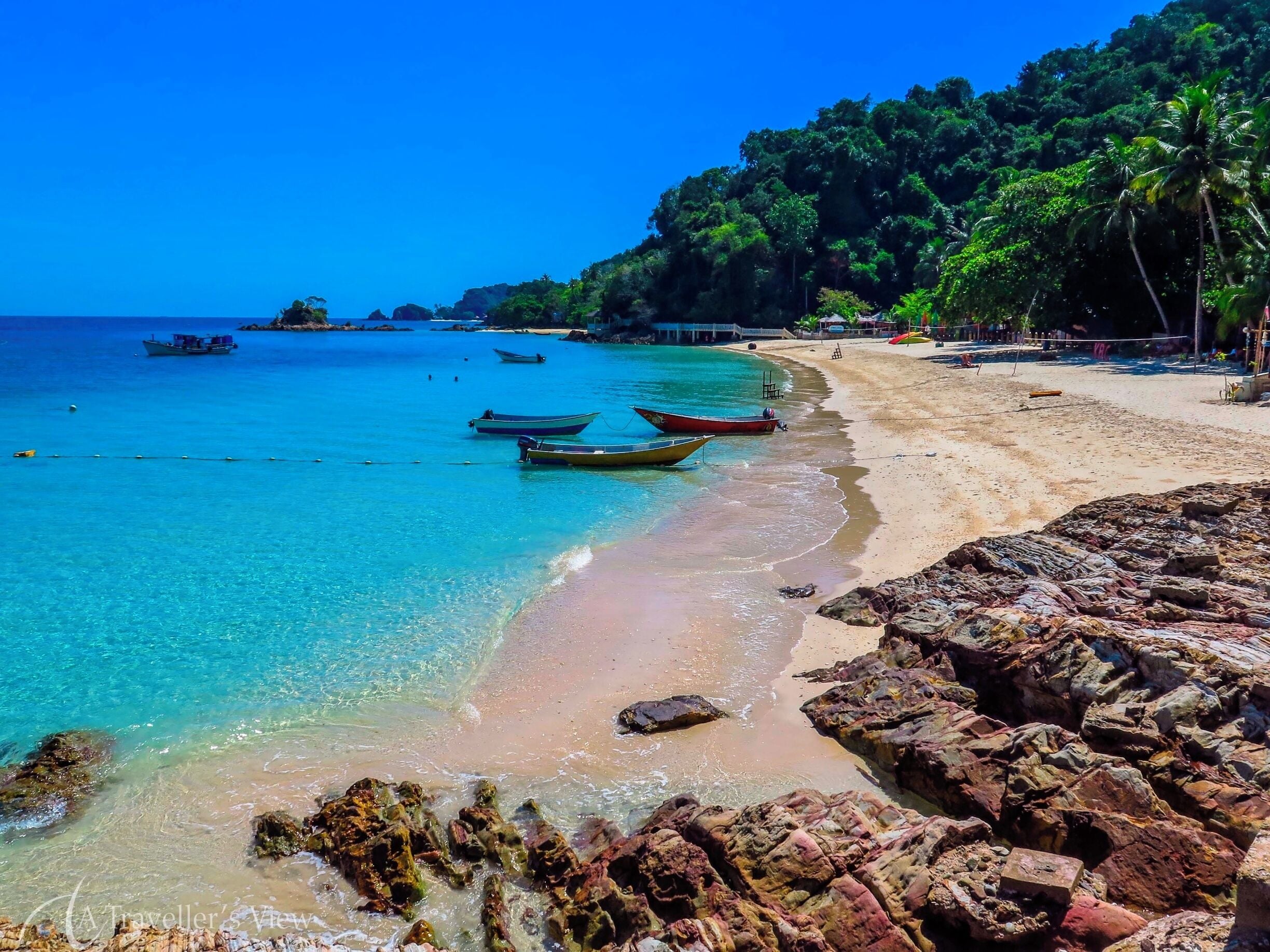 A beach with clear turquoise water, several boats anchored near the shore, and rocky outcrops.