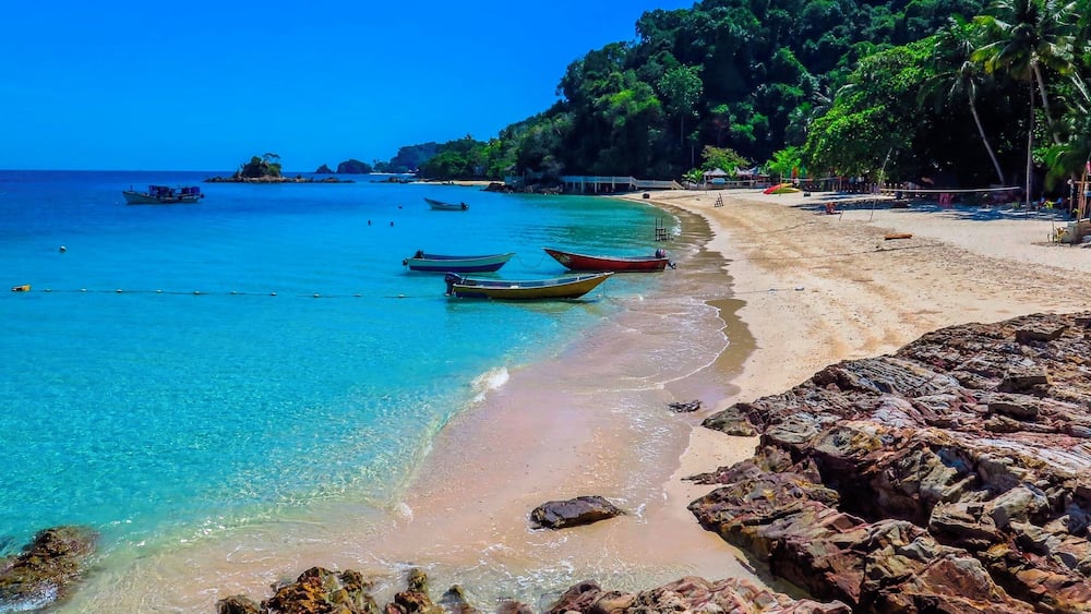 A beach with clear turquoise water, several boats anchored near the shore, and rocky outcrops.