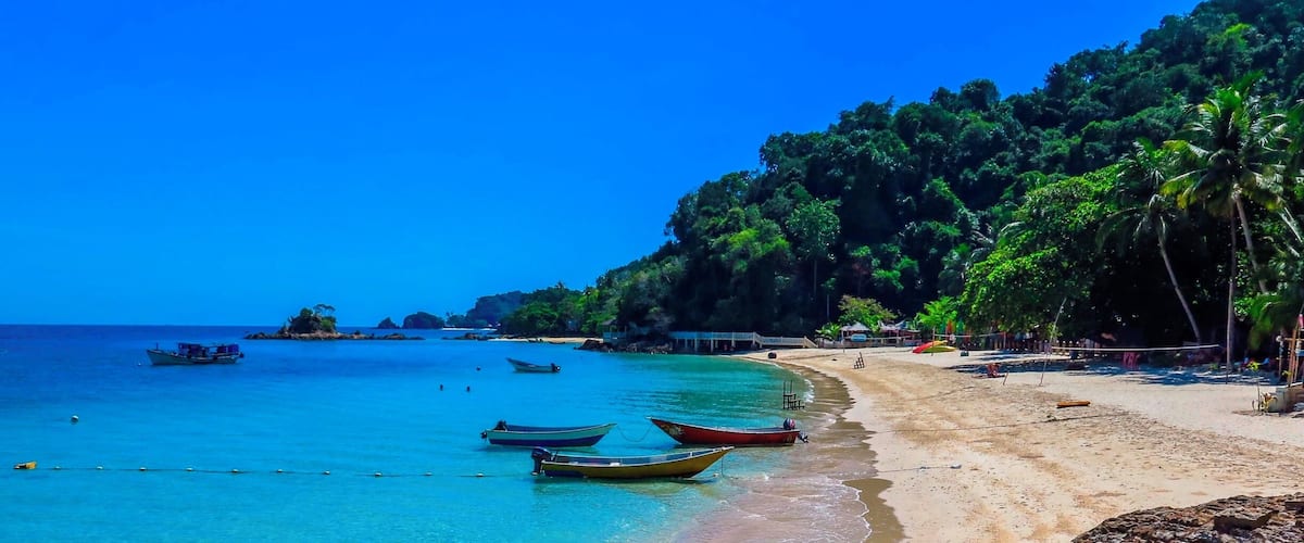 A beach with clear turquoise water, several boats anchored near the shore, and rocky outcrops.