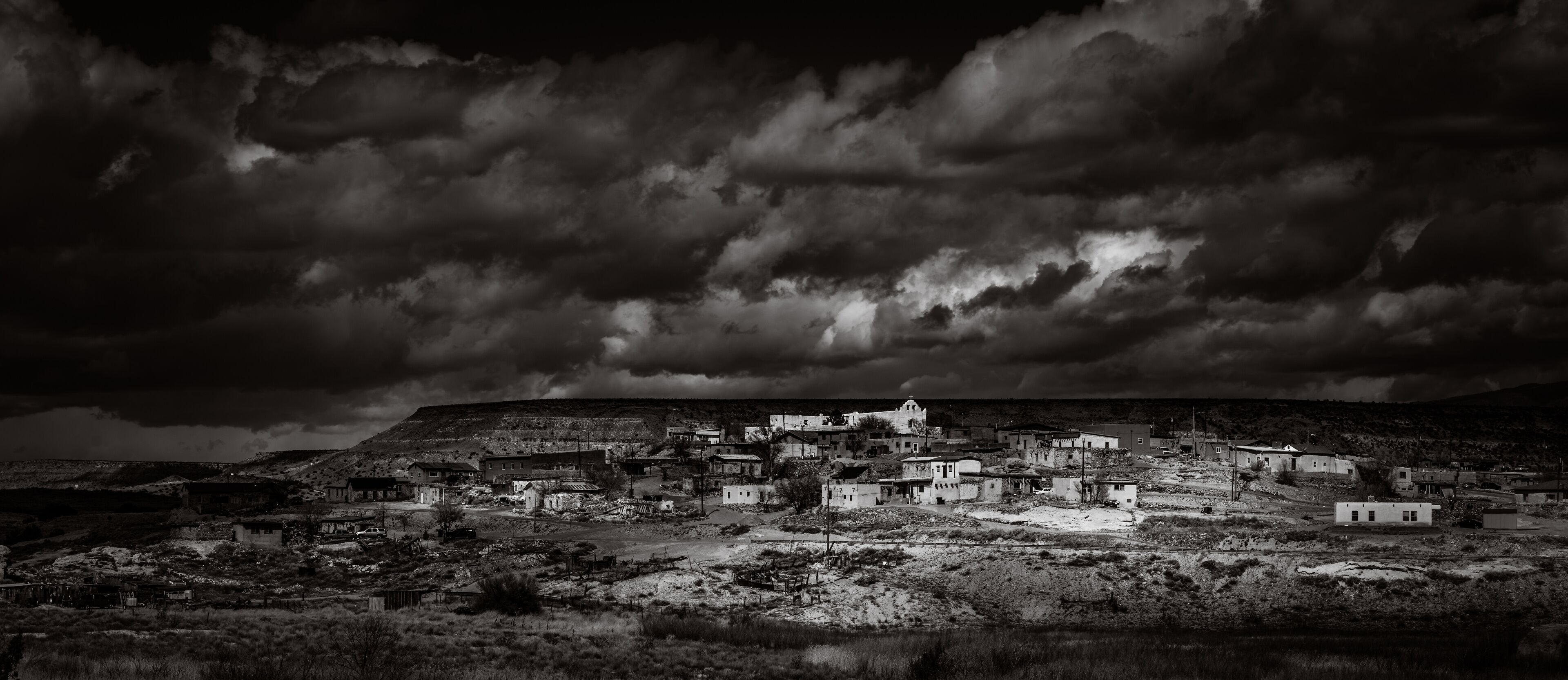 A black and white landscape of a small town with buildings and a hill in the background.