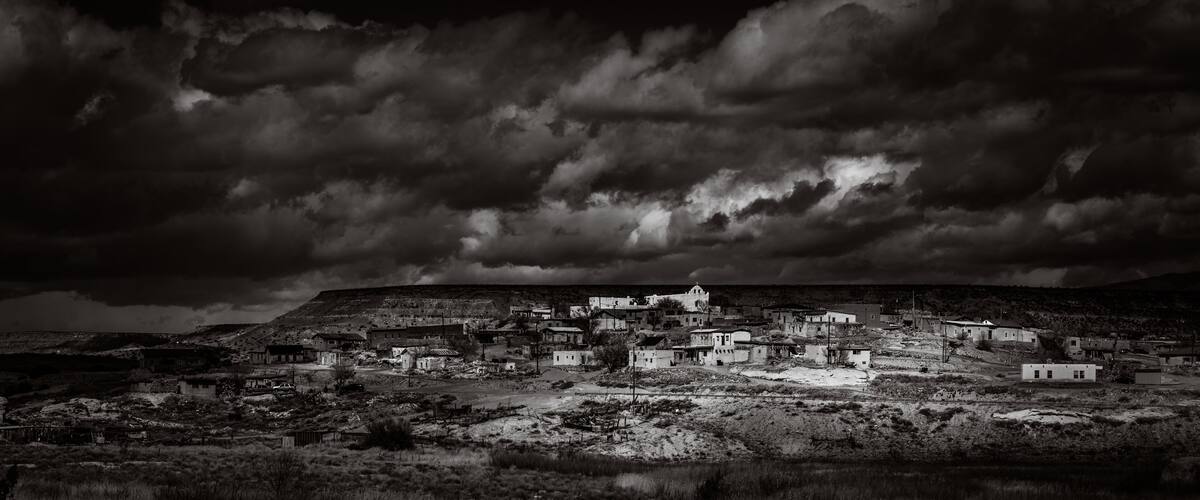 A black and white landscape of a small town with buildings and a hill in the background.