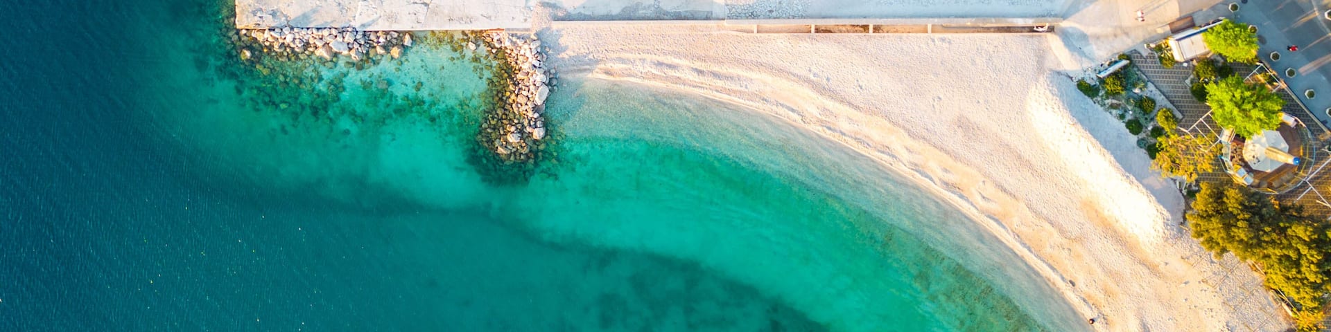 An aerial view of a coastal area with clear turquoise waters, a sandy beach, and a white pier extending into the water.