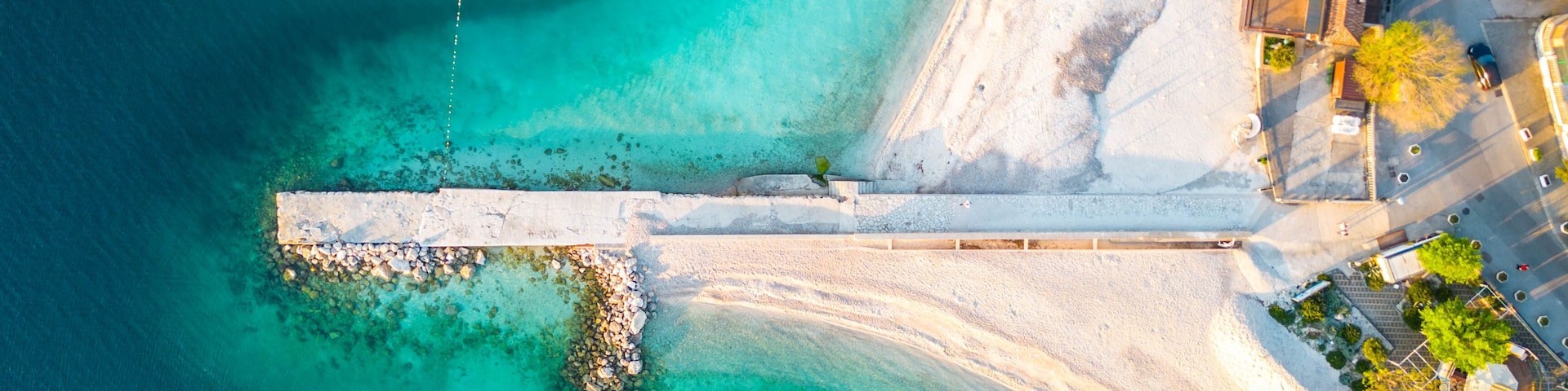 Luftaufnahme einer KĂŒstenlandschaft mit klarem tĂŒrkisblauem Wasser, einem Sandstrand und einem weiĂen Steg, der ins Wasser fĂŒhrt.