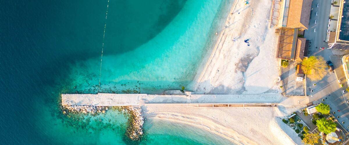 An aerial view of a coastal area with clear turquoise waters, a sandy beach, and a white pier extending into the water.