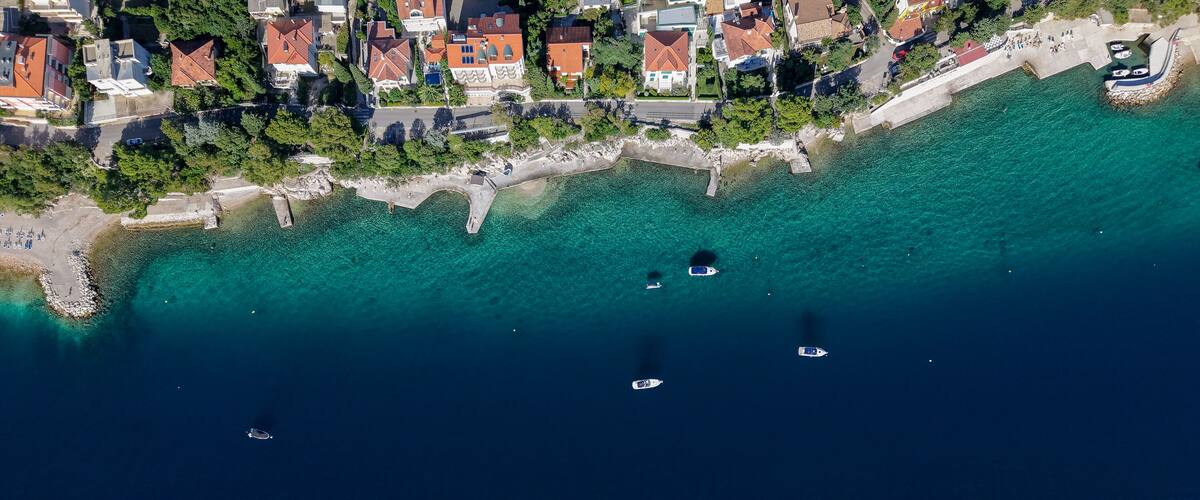 An aerial view of a coastal town with clear turquoise waters, boats, and houses with red roofs.