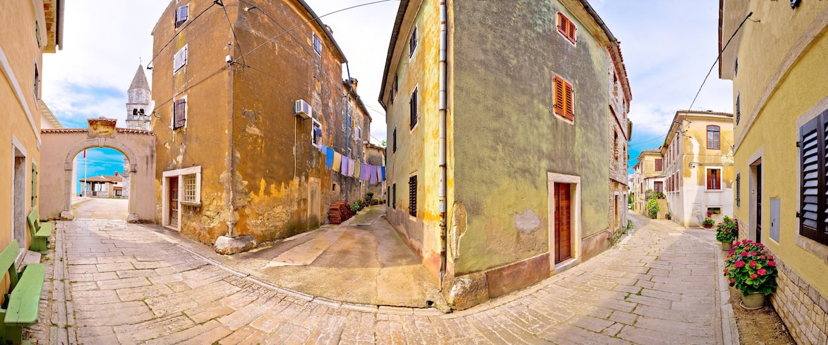 A narrow cobblestone street flanked by historic buildings with arched doorways and colorful shutters.