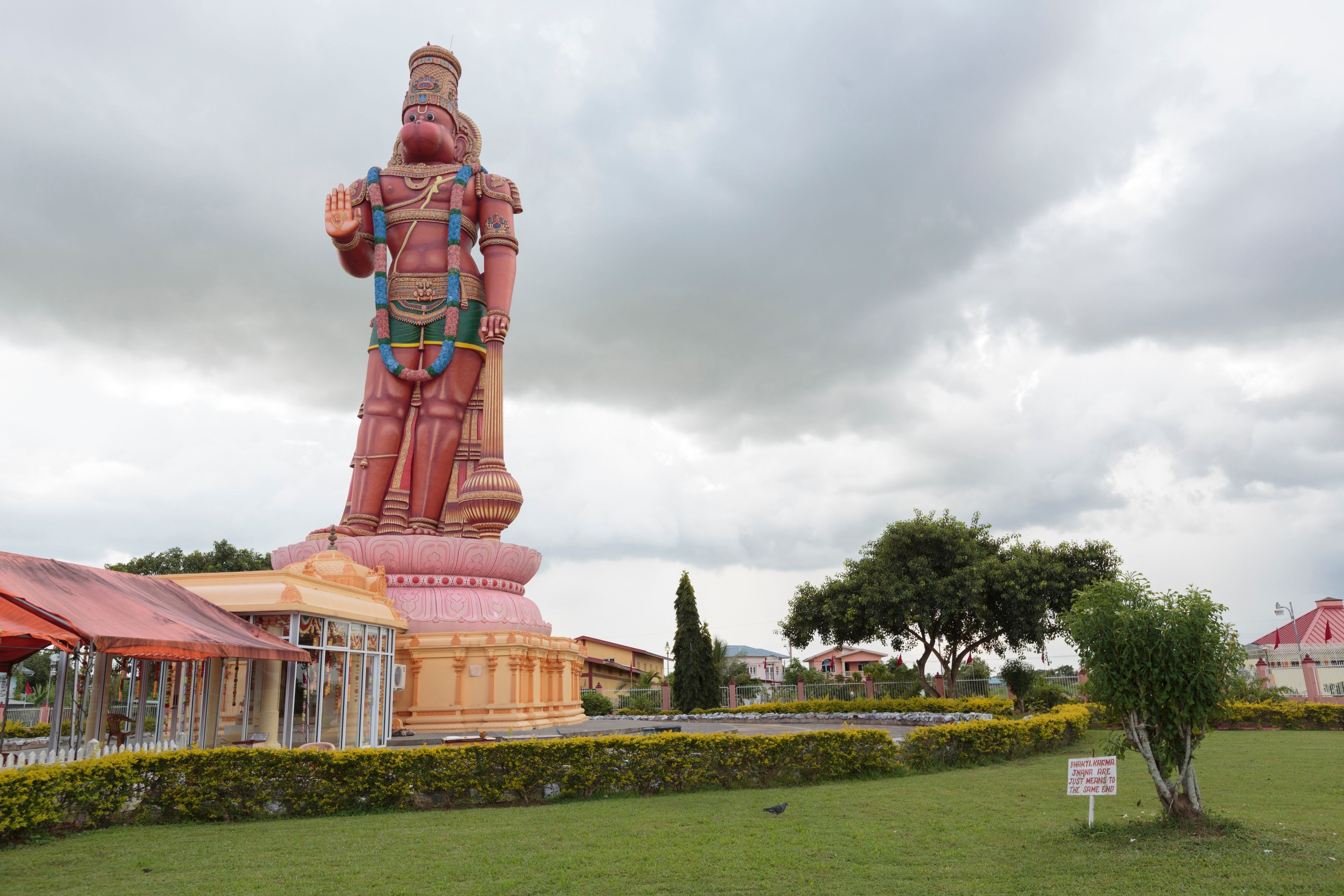 Eine große Statue einer Gottheit mit aufwendlichem Schmuck und einer traditionellen Kopfbedeckung, umgeben von einem gepflegten Garten und einem kleinen Pavillon.