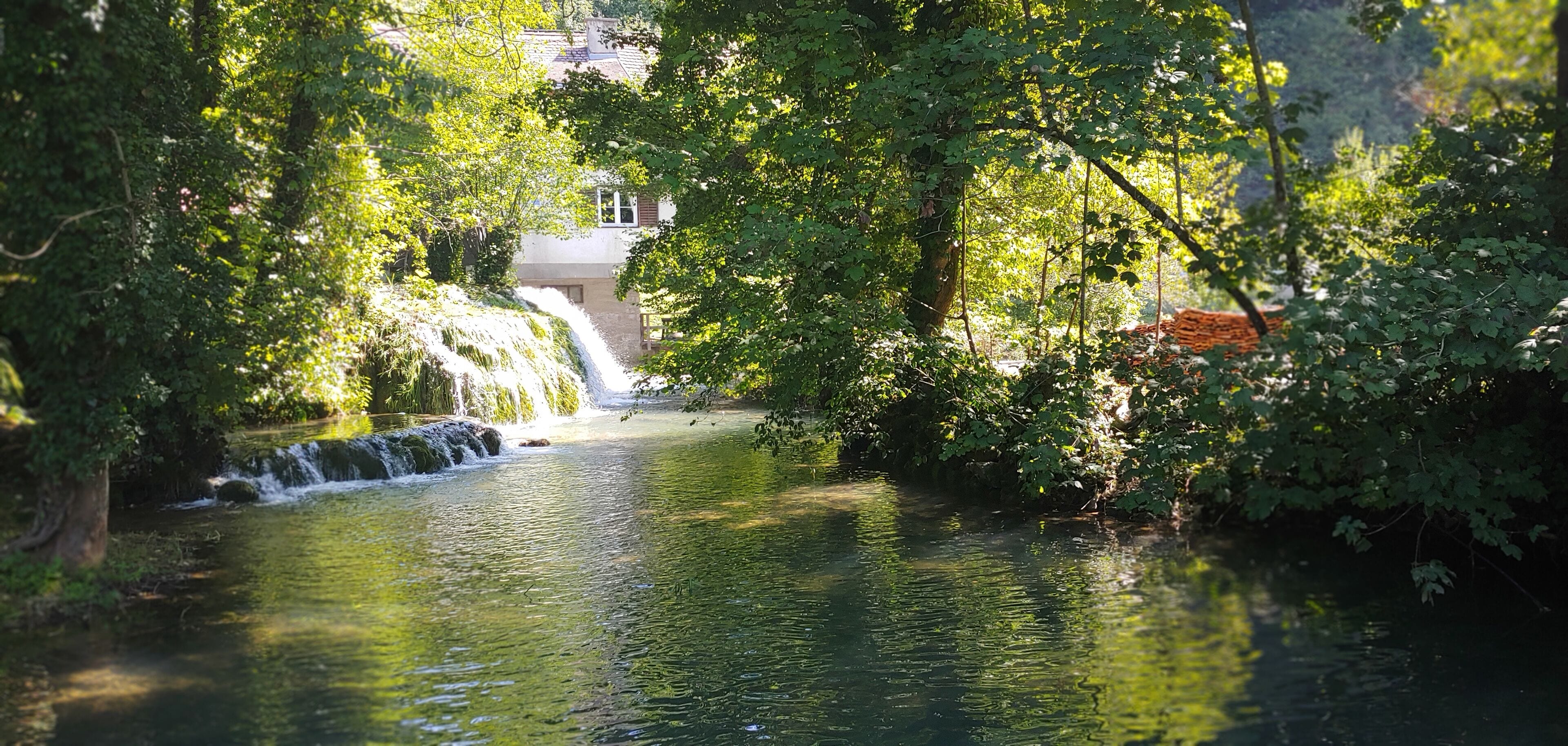 A serene river with a small waterfall, surrounded by lush greenery and trees.