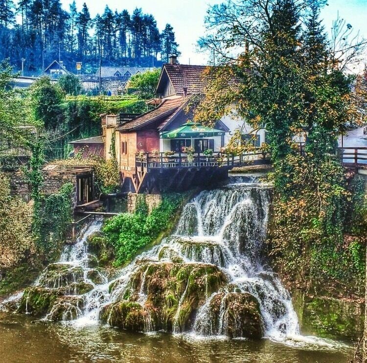 A waterfall cascading over moss-covered rocks, surrounded by lush greenery and traditional buildings.