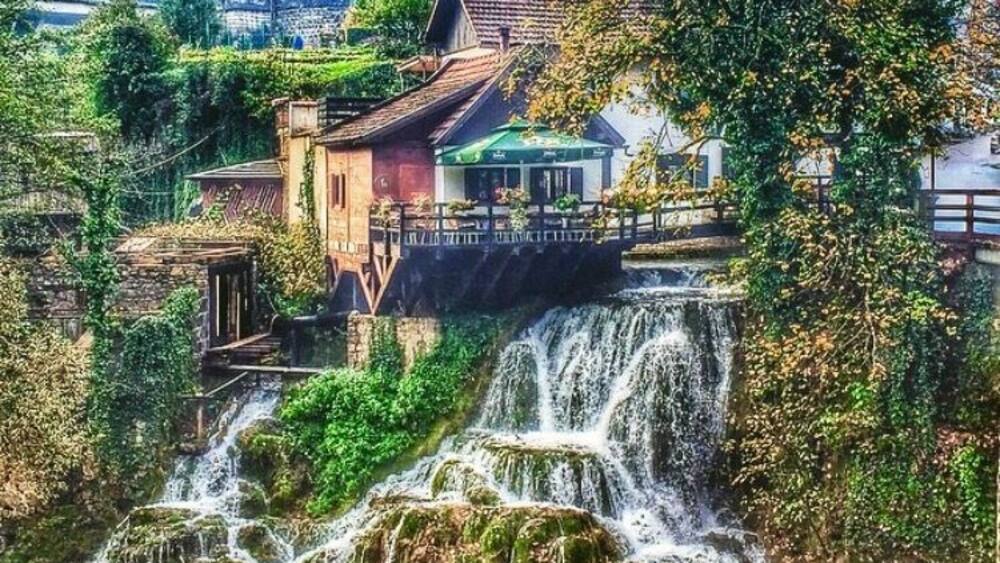 A waterfall cascading over moss-covered rocks, surrounded by lush greenery and traditional buildings.