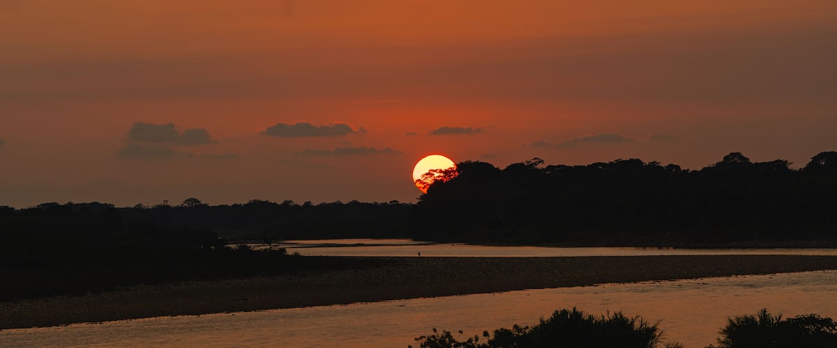 A sunset over a river with silhouettes of trees and a distant horizon.