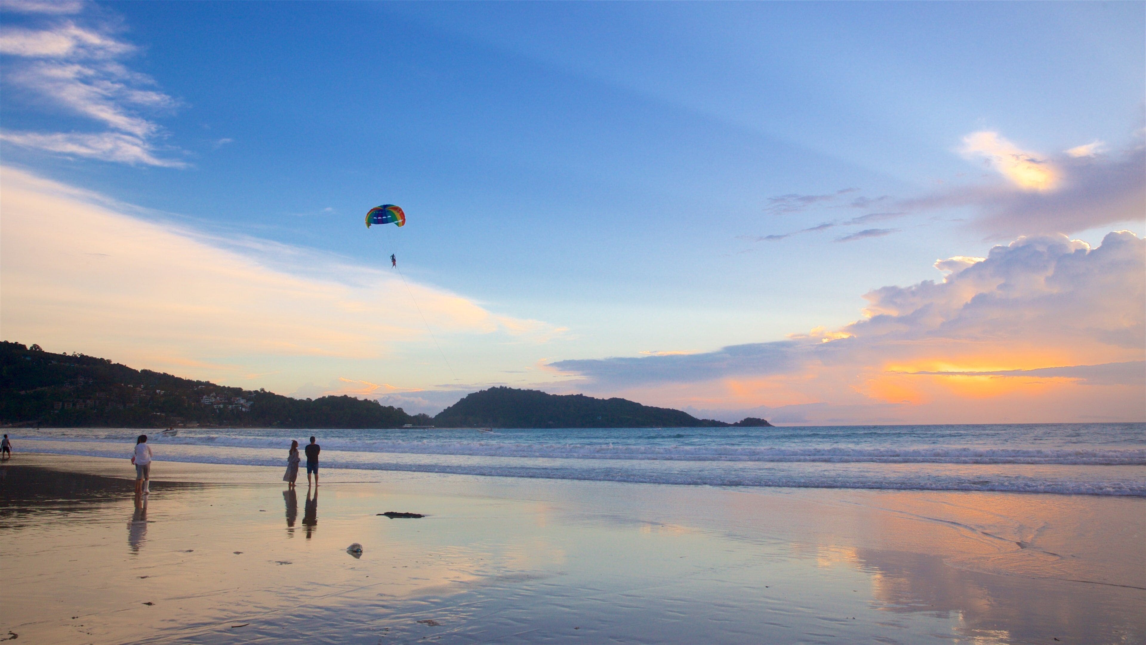 Una playa con gente, un kitesurfista y un atardecer sobre el océano.