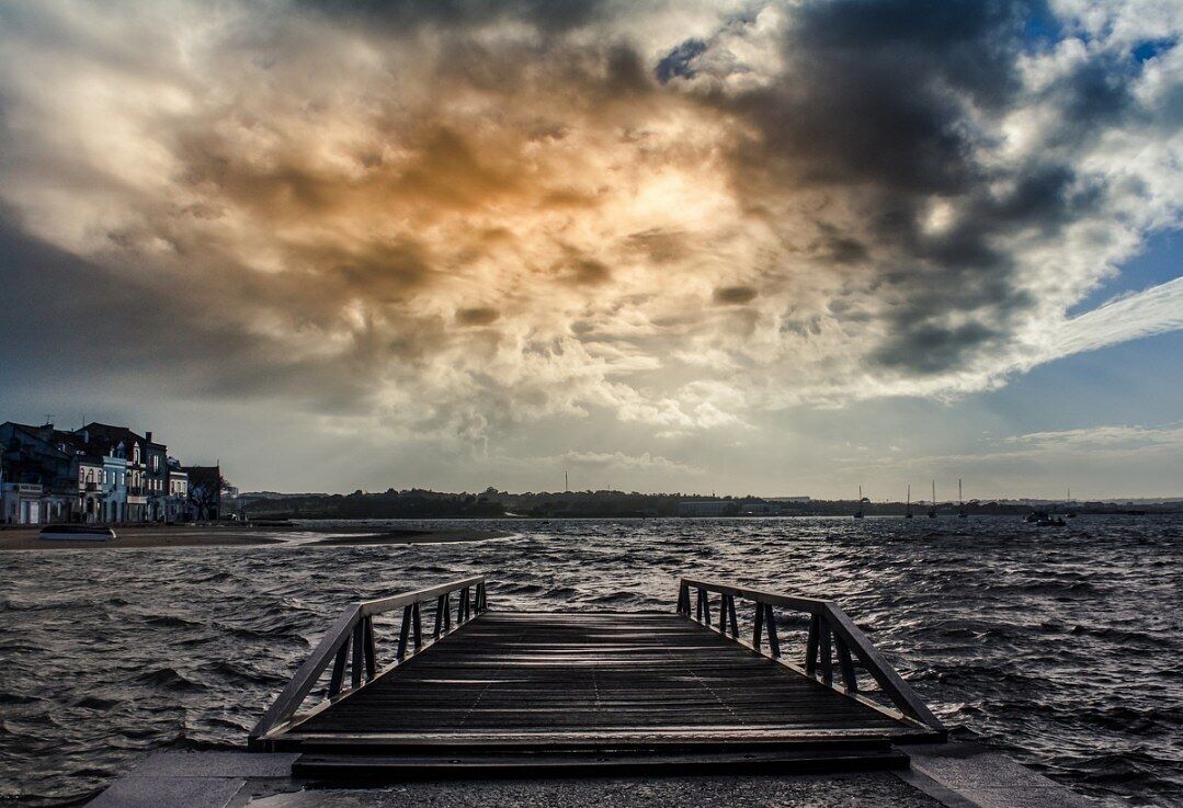 A wooden pier leading to a beach with buildings in the background under a cloudy sky.
