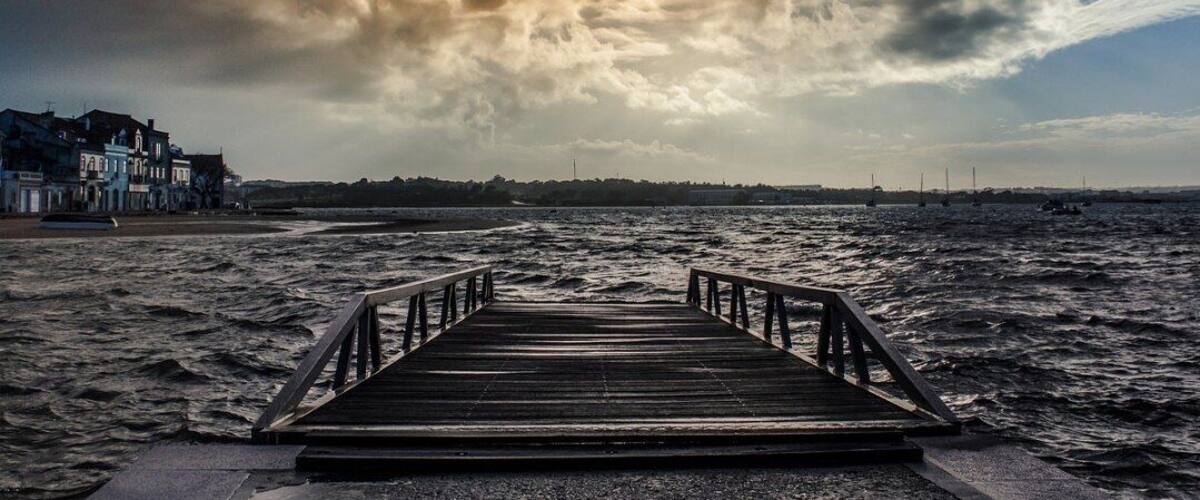 A wooden pier leading to a beach with buildings in the background under a cloudy sky.