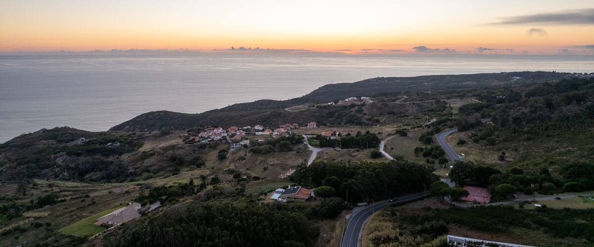 A coastal landscape with a winding road, a small settlement, and a view of the sea at sunset.