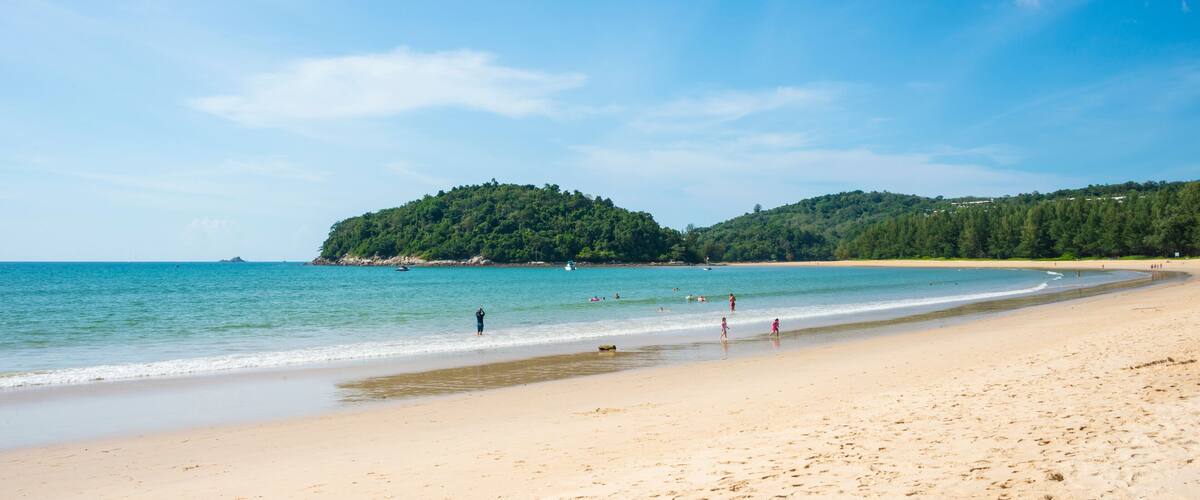 A sandy beach with clear blue water, a small island in the distance, and a few people enjoying the shore.