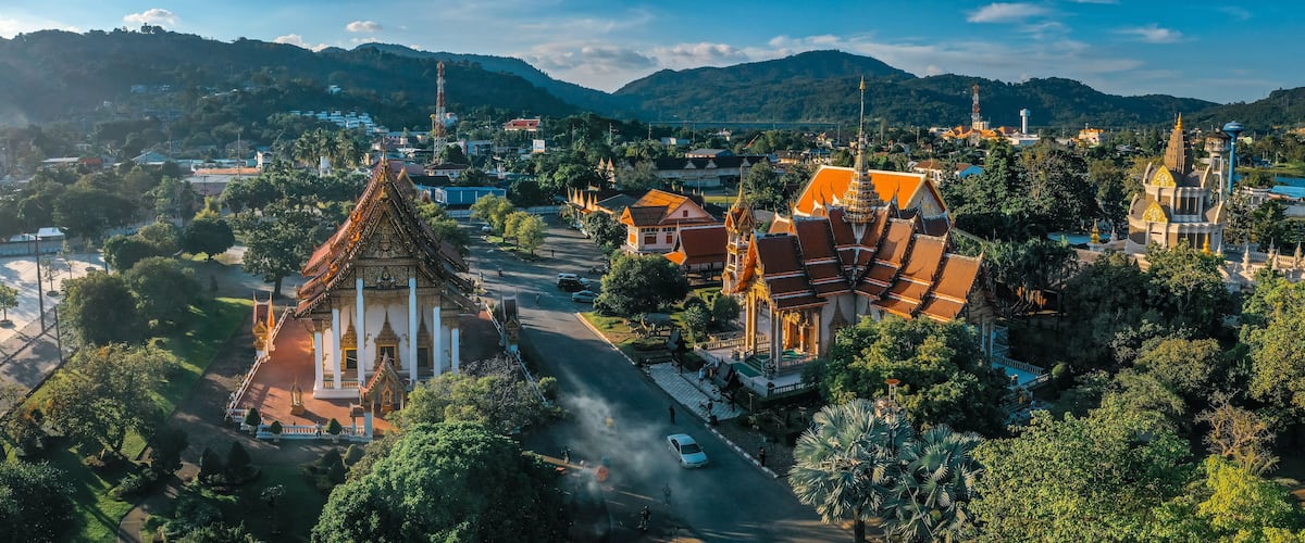 A temple complex with traditional architecture, surrounded by lush greenery and a mountainous backdrop.
