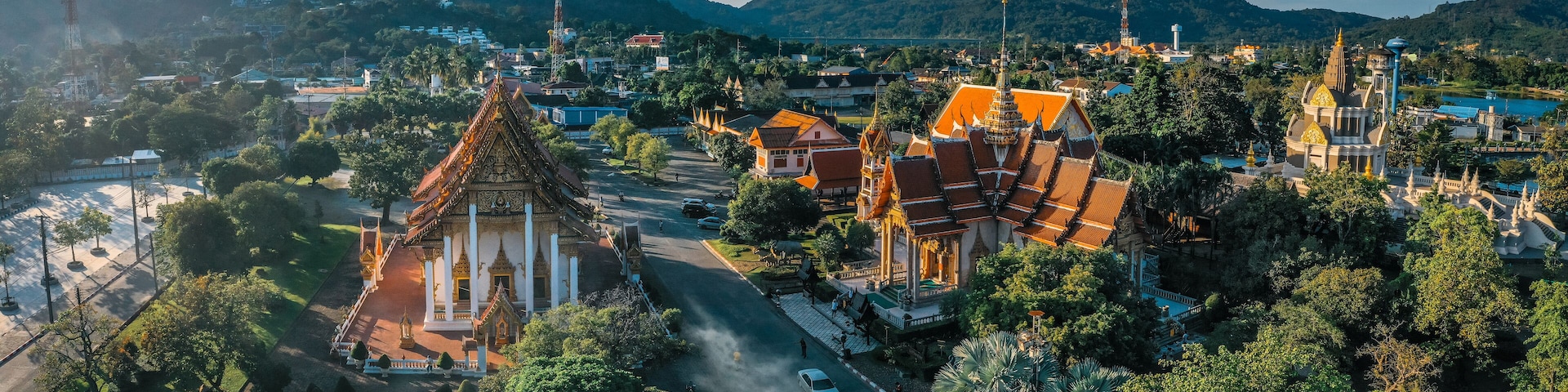 A temple complex with traditional architecture, surrounded by lush greenery and a mountainous backdrop.