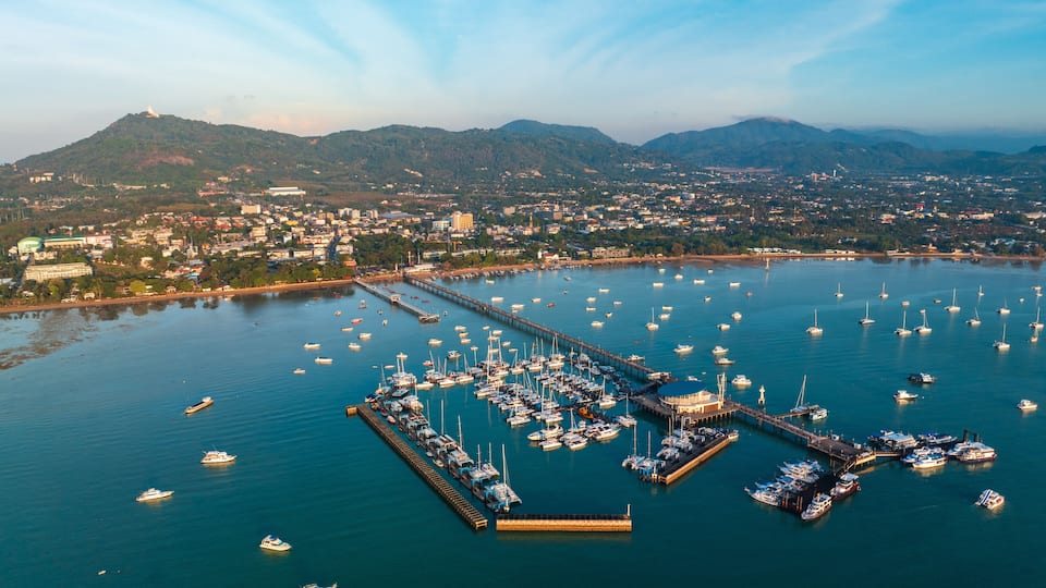 A marina with numerous boats docked, surrounded by a coastal town and mountains in the background.