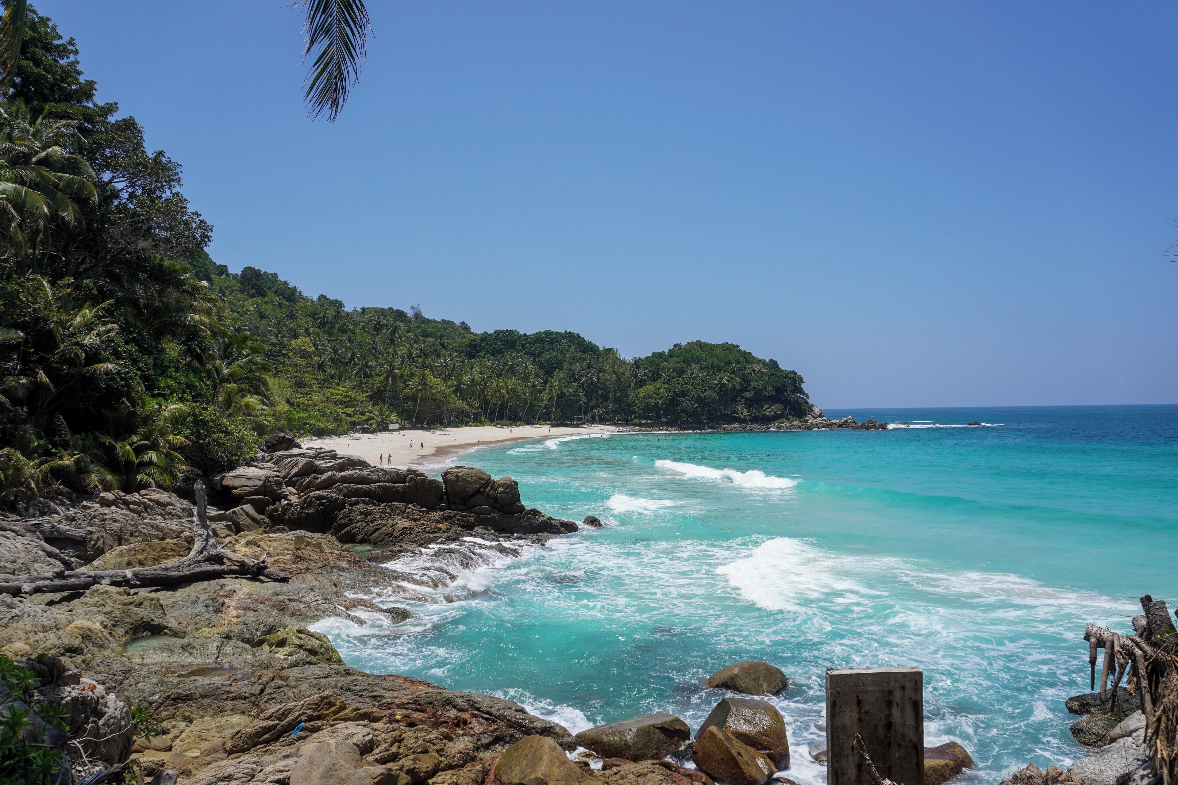 A tropical beach with clear turquoise waters, rocky shore, and dense green foliage.