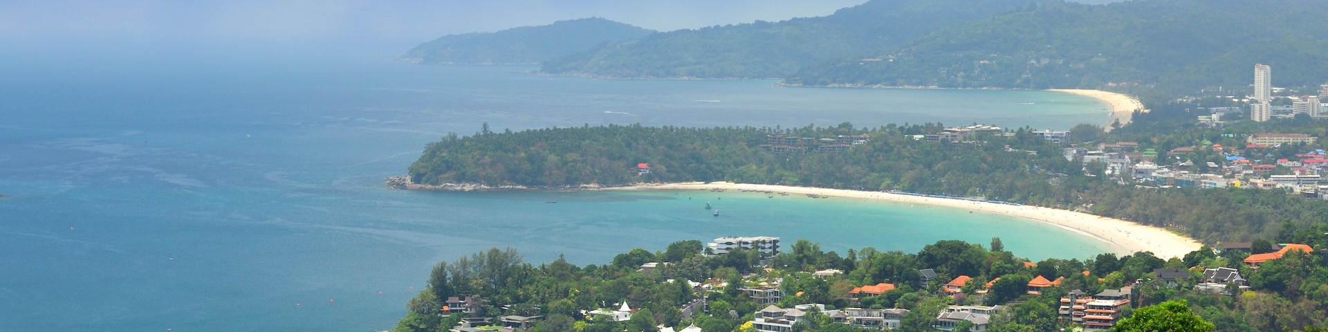 Paisagem costeira com uma praia arenosa, águas azul-turquesa e uma colina arborizada.