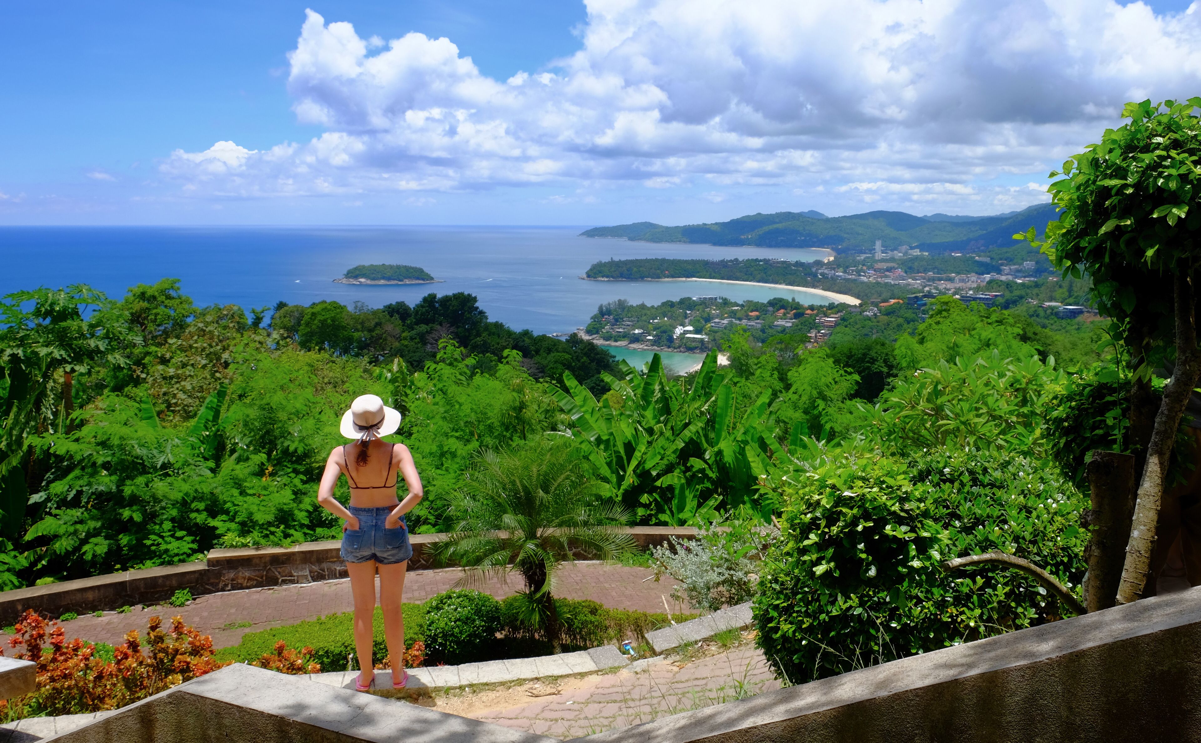 A woman standing on a stone ledge overlooking a coastal landscape with lush greenery and a view of the sea and islands.