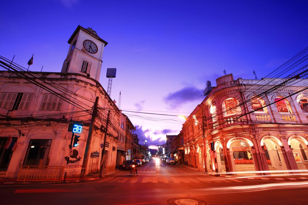 A street at dusk with a clock tower, illuminated buildings, and overhead power lines.