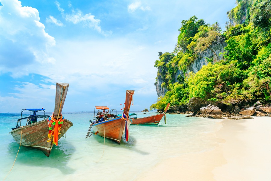 Twee traditionele houten boten met kleurrijke decoraties aan een zandstrand, met helder turquoise water en groene kliffen op de achtergrond.