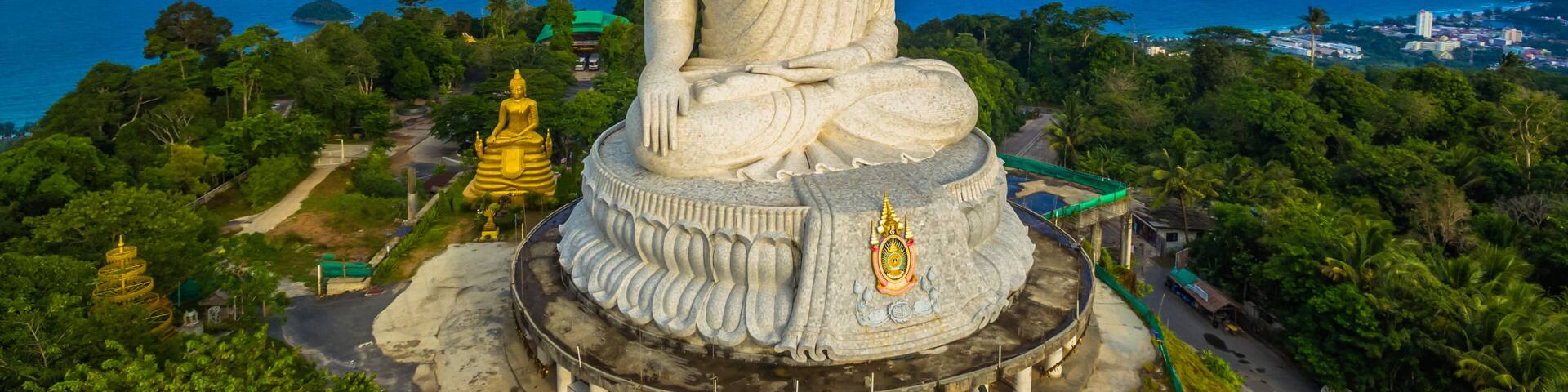 A large white Buddha statue with a golden base, surrounded by lush greenery and overlooking a coastal landscape.