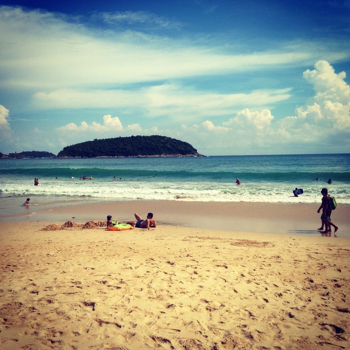 A sandy beach with footprints, a distant forested island, and people in the water.
