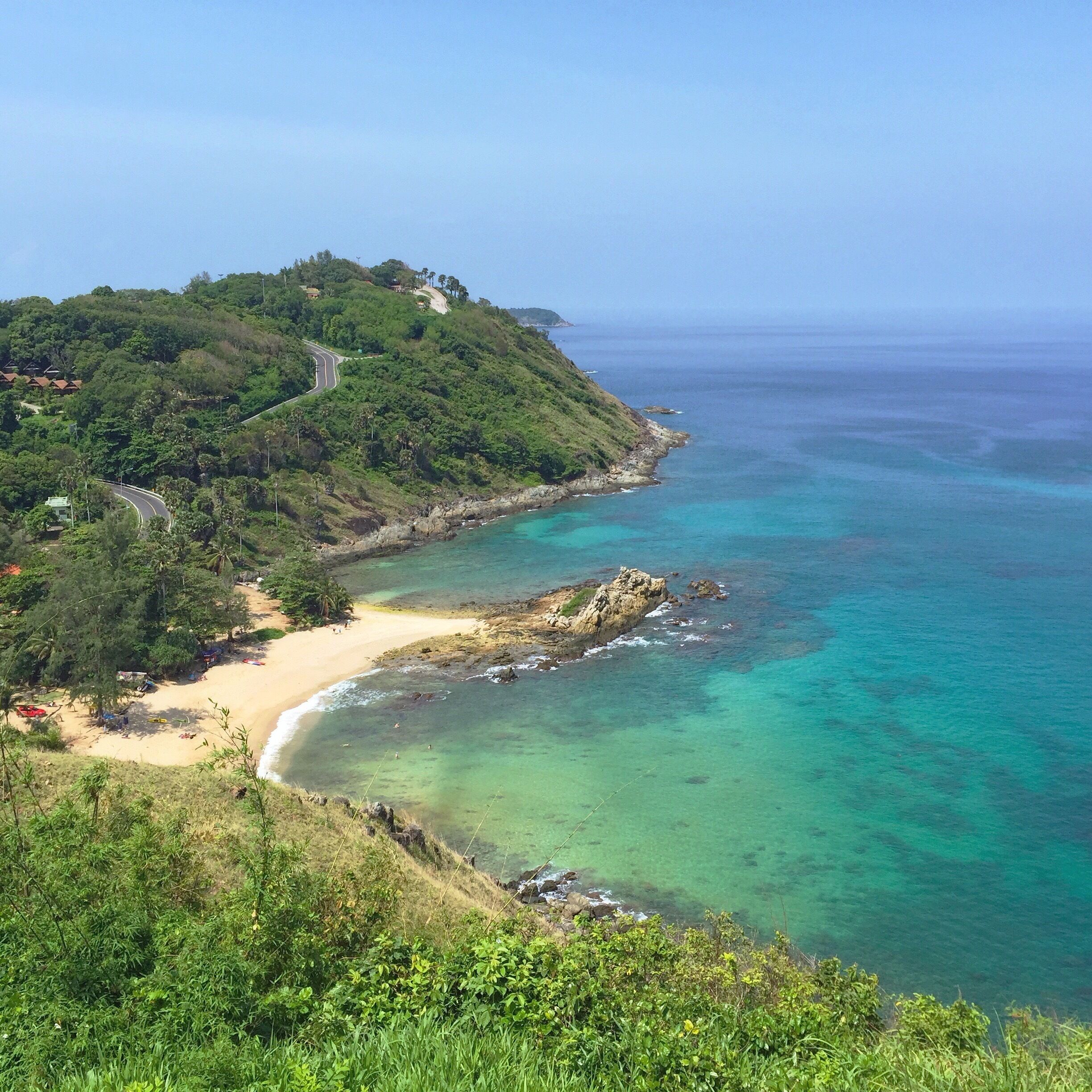 A coastal landscape with a sandy beach, clear turquoise waters, and a lush green hill with a winding road.