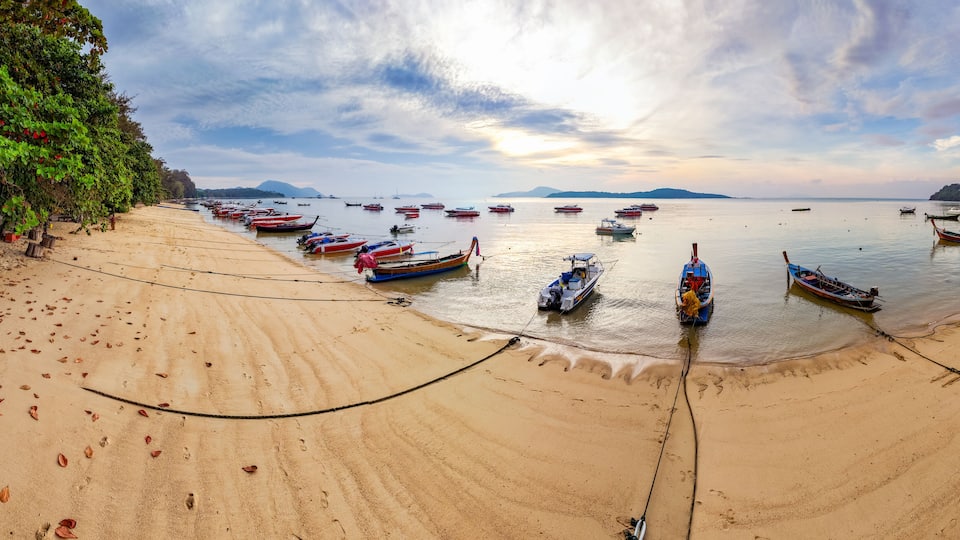 A beach with numerous boats anchored in calm waters, a sandy shore, and a backdrop of distant mountains.
