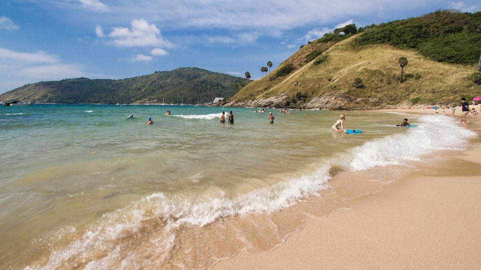 A beach with clear turquoise water, people swimming and surfing, and a hilly landscape in the background.