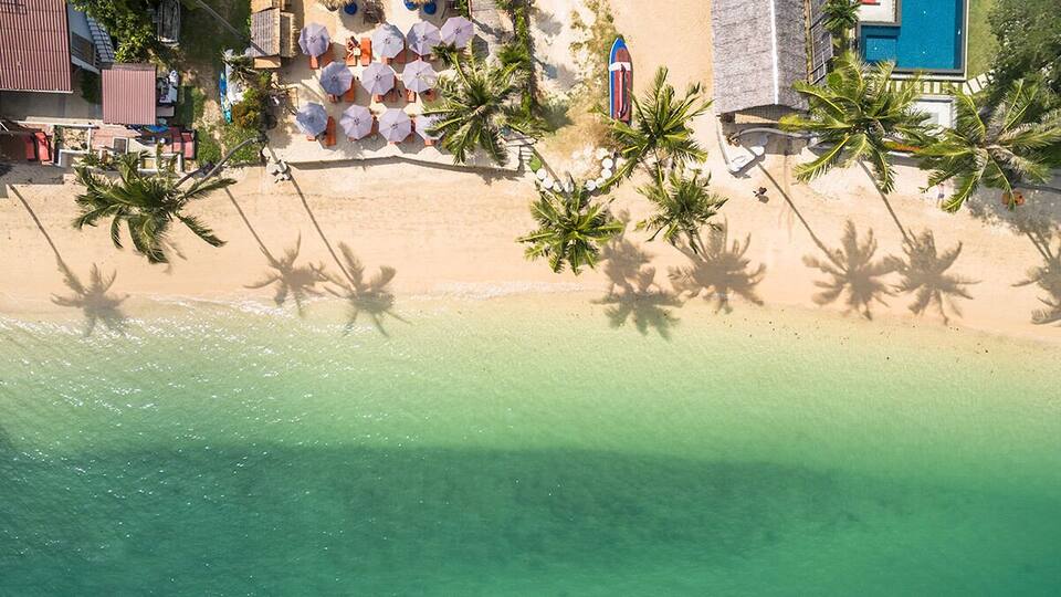 A beach with clear turquoise water, palm trees, and a resort area with umbrellas and lounge chairs.