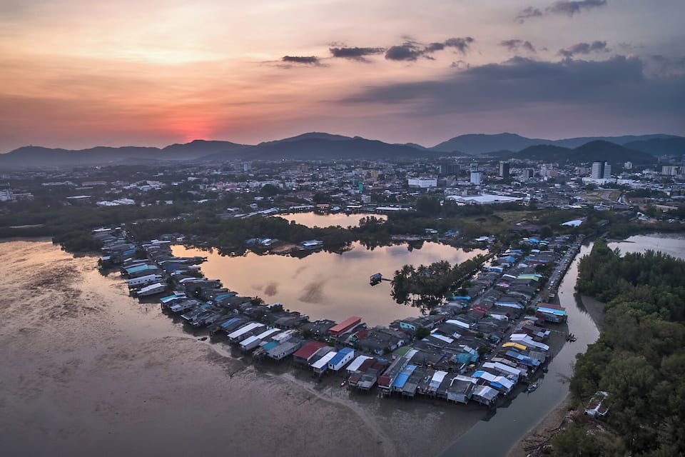 An aerial view of a coastal city with a heart-shaped cluster of buildings near a body of water, surrounded by mountains and a sunset sky.