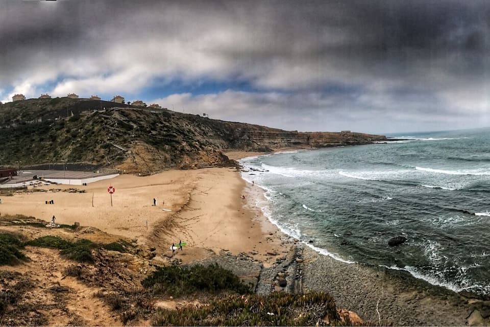 Paisagem costeira com praia arenosa, falésias rochosas e céu tempestuoso.