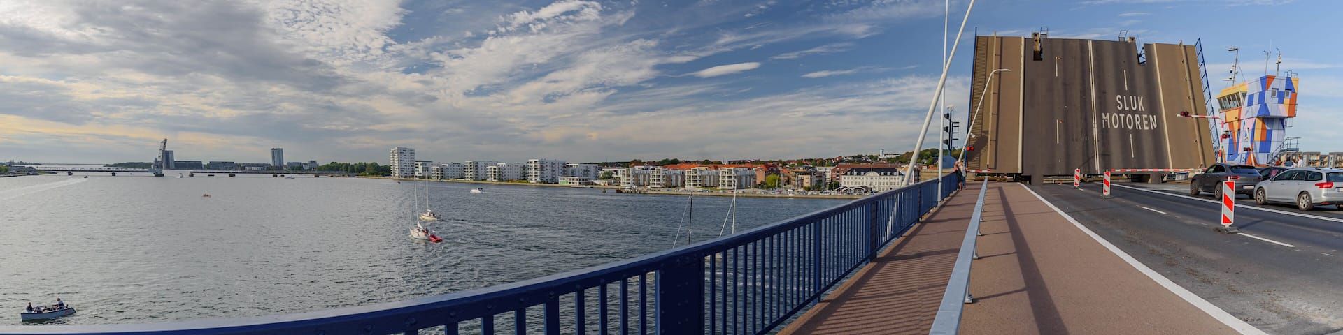 A bridge with a blue railing, a river, sailboats, and buildings in the background.