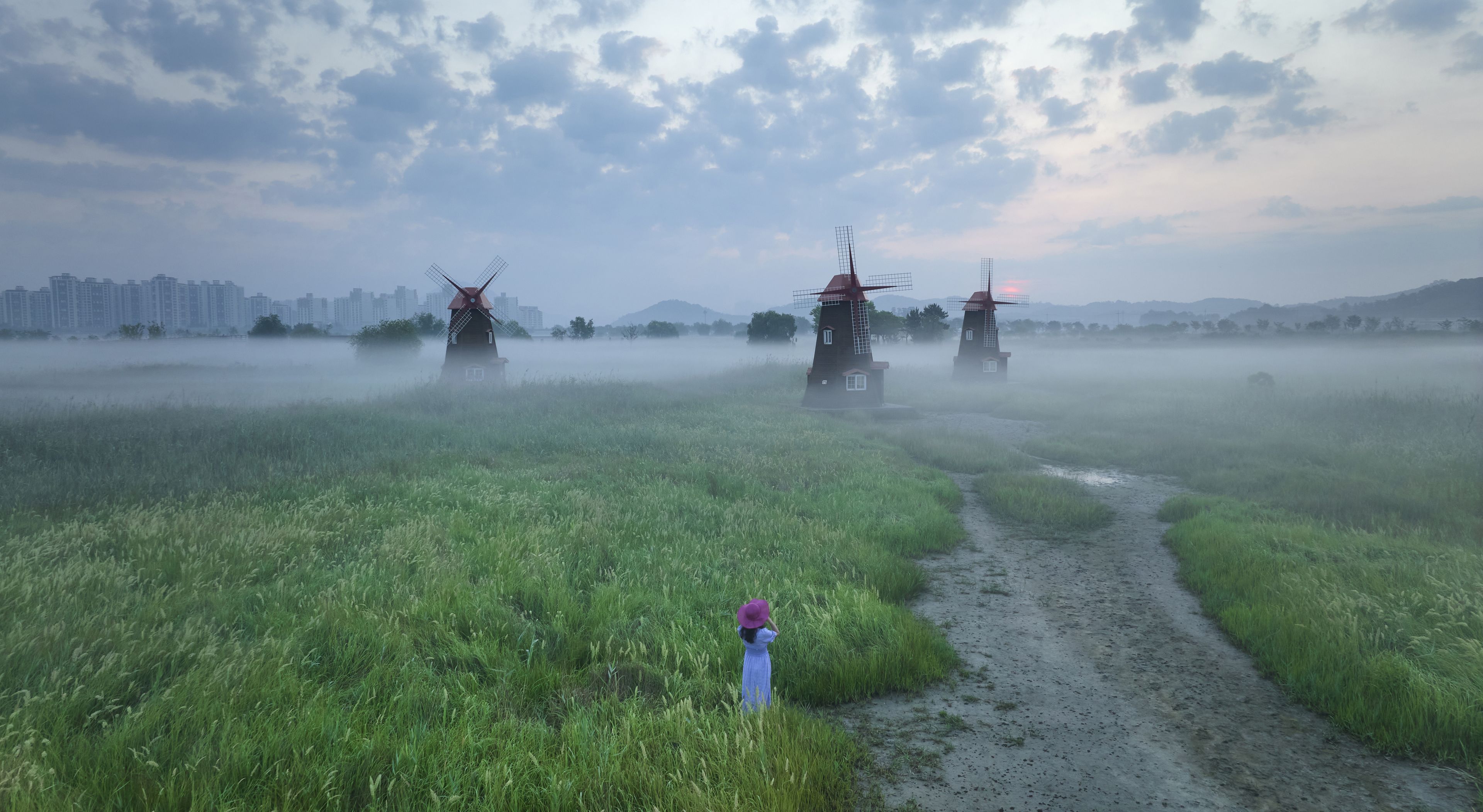 Namdong-gu, Incheon, South Korea - July 3, 2022: Aerial and morning view of a female in red hat standing on ground against windmills with fog at Sorae Wetland Ecological Park in summer