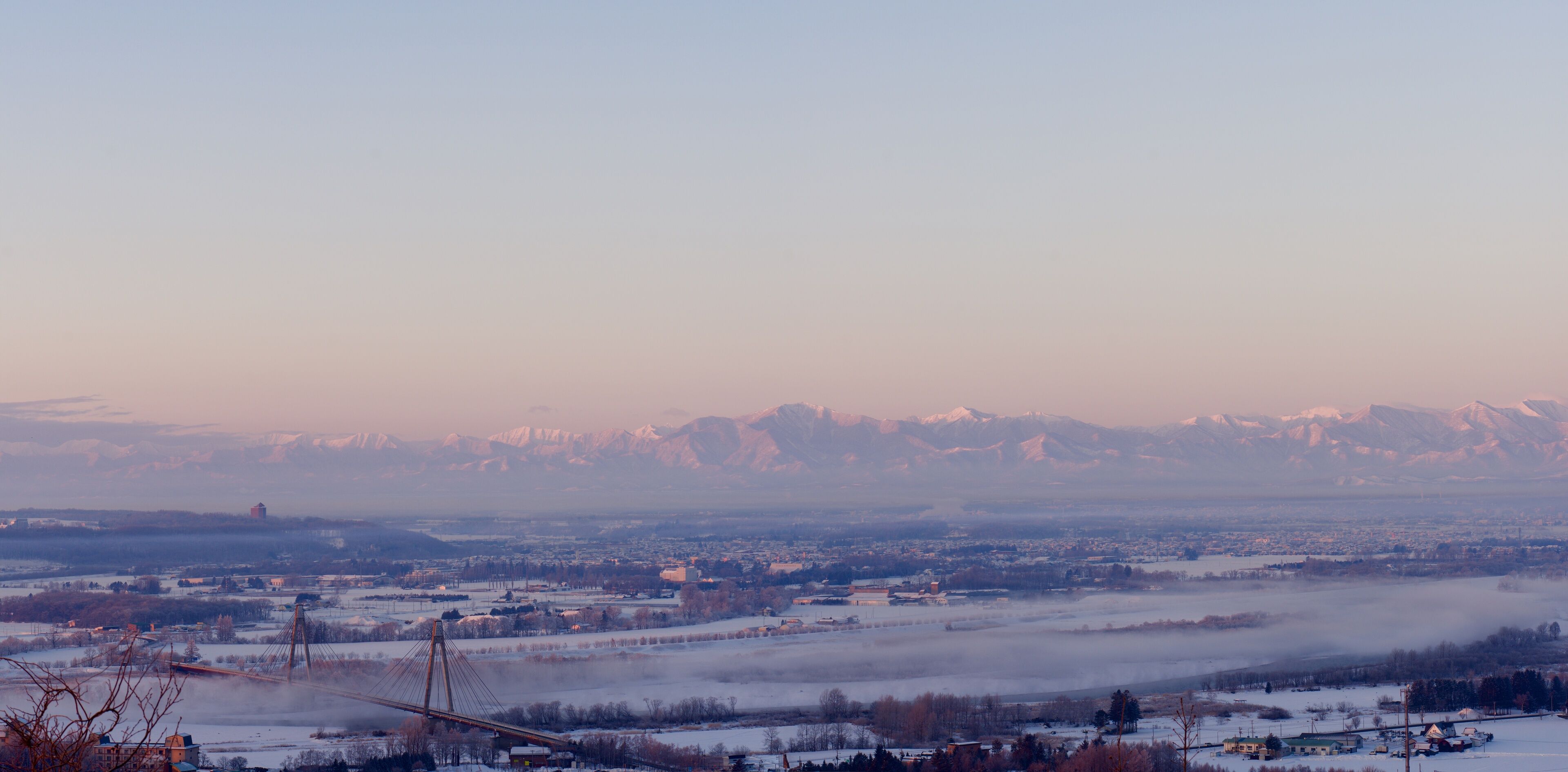 A snow-covered landscape with a bridge, mountains, and a town in the background.